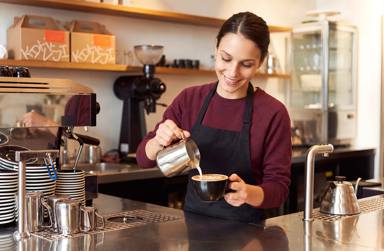 A Kahveji barista in a plum sweater pouring steamed milk into a cup behind the café counter, with warm-lit shelves of Kahveji kraft boxes and ceramic cups behind her.